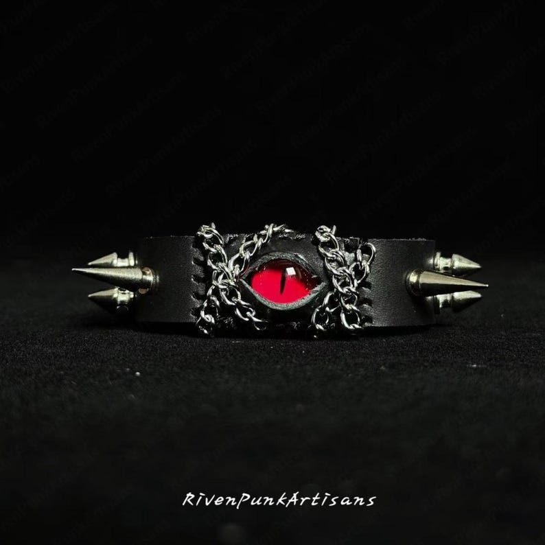May include: Black leather cuff bracelet featuring a red eye design with silver chain accents. The bracelet has silver cone studs on each end. The text "RivenPunkArtisans" is at the bottom.