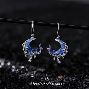 May include: A pair of silver-toned earrings featuring a crescent moon design with a blue enamel inlay. The earrings have a dripping, textured silver border and a dangling hook. The text "RivenPunkArtisans" is at the bottom.