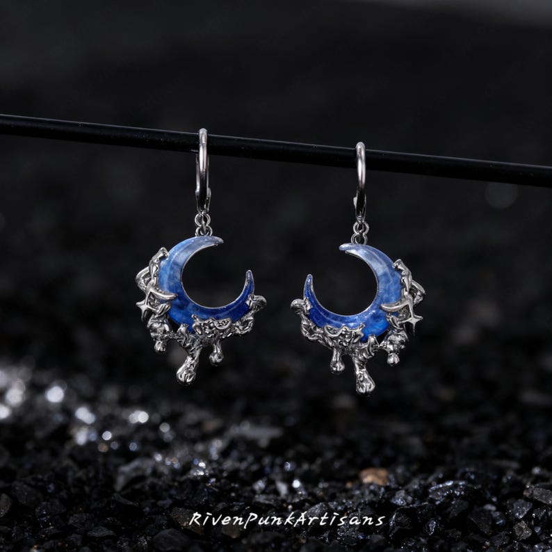 May include: A pair of silver-toned earrings featuring a crescent moon design with a blue enamel inlay. The earrings have a dripping, textured silver border and a dangling hook. The text "RivenPunkArtisans" is at the bottom.