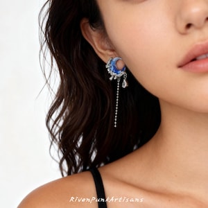 May include: A close-up of a blue and silver crescent moon earring with a dangling chain and a teardrop-shaped charm. The earring has intricate detailing and is worn on the ear. The text "RivenPunkArtisans" is at the bottom.