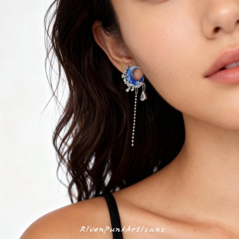 May include: A close-up of a blue and silver crescent moon earring with a dangling chain and a teardrop-shaped charm. The earring has intricate detailing and is worn on the ear. The text "RivenPunkArtisans" is at the bottom.