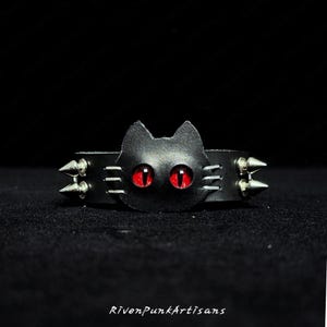 Może przedstawiać: Czarna skórzana bransoletka z motywem kociej twarzy, czerwonymi oczami i srebrnymi kolcami. Twarz kota jest centralna, z trzema liniami po każdej stronie. Wyjątkowy dodatek.