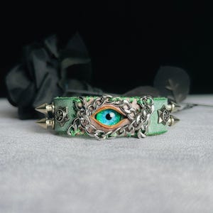 May include: A green leather bracelet with a silver chain and a blue eye design. The bracelet has silver spikes on either side.
