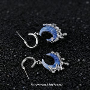May include: A pair of silver-toned earrings featuring a crescent moon design with a blue and white marbled effect. The earrings have a detailed, textured silver accent and a post-and-hoop closure. The text "RivenPunkArtisans" is at the bottom.