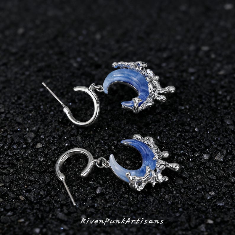 May include: A pair of silver-toned earrings featuring a crescent moon design with a blue and white marbled effect. The earrings have a detailed, textured silver accent and a post-and-hoop closure. The text "RivenPunkArtisans" is at the bottom.