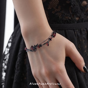 May include: A black and red bracelet with a gothic theme. The bracelet features a detailed raven design, red gemstone accents, and a delicate chain. The bracelet is worn on a wrist, with the text "RivenPunkArtisans" visible at the bottom.