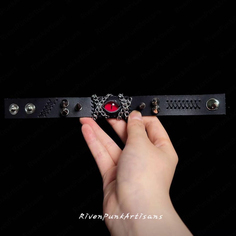 May include: Black leather bracelet featuring a central red eye design framed by silver-colored chains. The bracelet is adorned with silver studs, rivets, and decorative stitching. The text "RivenPunkArtisans" is visible at the bottom.