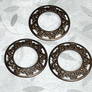 May include: Three bronze-colored, open-center metal rings with ornate, floral-inspired filigree designs. The rings are circular, with a detailed pattern around the outer and inner edges. The background is a light gray with a subtle pattern.