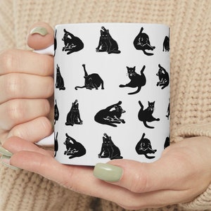 May include: White ceramic mug with a black and white pattern of cats in various poses. The mug is being held by a person's hands.