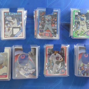 May include: Baseball trading cards featuring Chicago Cubs players, displayed in protective sleeves. Cards include Sammy Sosa, Trey Mancini, and others. The cards are secured with blue tape and are on a blue surface.