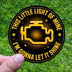 May include: A round black sticker with the text "THIS LITTLE LIGHT OF MINE I'M GONNA LET IT SHINE" in yellow. The sticker features a yellow engine light icon in the center, with star accents. The sticker is held by a hand against a green grass background.