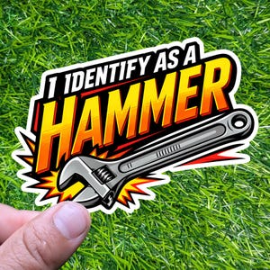 May include: A sticker with the text "I IDENTIFY AS A HAMMER" in white, yellow, and orange, with a gray adjustable wrench graphic. The sticker has a white border and a burst design, held by a hand against a green grass background.
