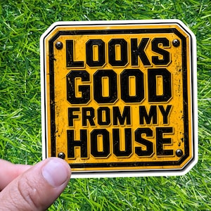 May include: A yellow and black sticker with the text "LOOKS GOOD FROM MY HOUSE" in bold, block letters. The sticker has a distressed, vintage look and is held against a green, grassy background.