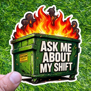 May include: A sticker featuring a green dumpster engulfed in flames. The text "ASK ME ABOUT MY SHIFT" is prominently displayed. A yellow "CAUTION" sign is on the side. The sticker is on a green, grassy background.