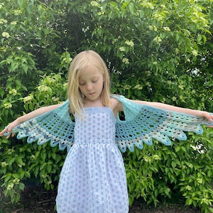 May include: A young girl wearing a blue and white dress with a crocheted blue and white shawl. The shawl is shaped like wings and the girl is holding her arms out to the sides.