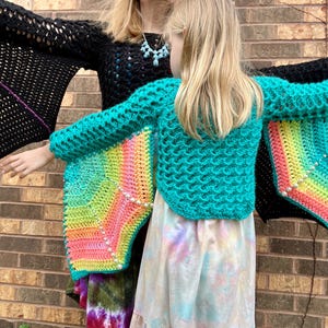 May include: Two people wearing crocheted sweaters with rainbow wings. The sweaters are teal and black with colorful crocheted wings.