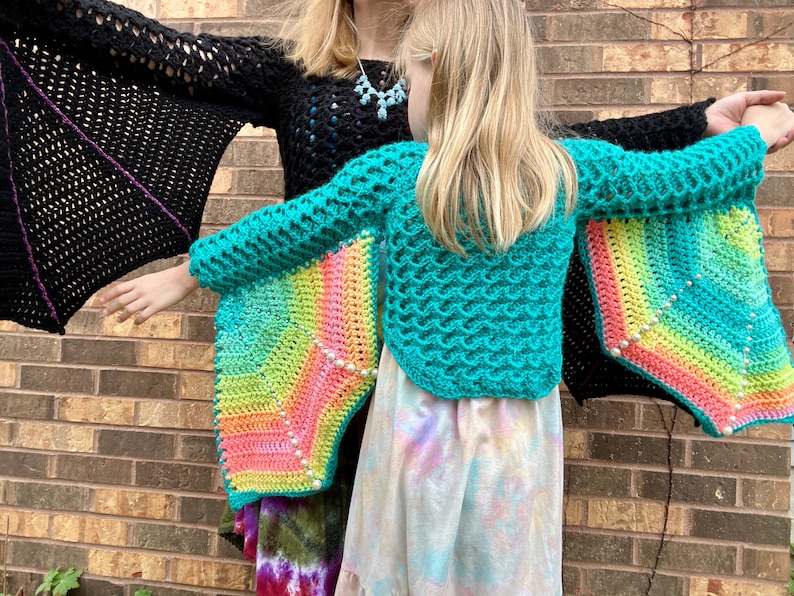 May include: Two people wearing crocheted sweaters with rainbow wings. The sweaters are teal and black with colorful crocheted wings.