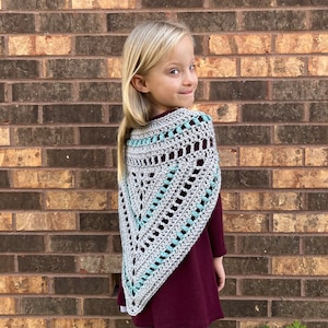 Puede incluir: Un chal de crochet gris y turquesa con forma triangular. El chal lo lleva puesto una persona joven con el pelo rubio.