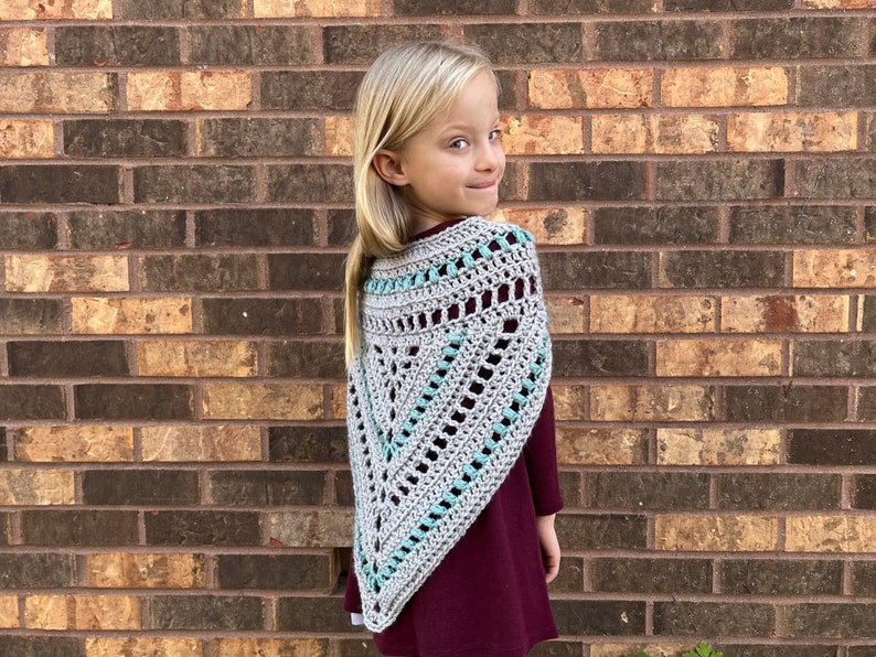 Puede incluir: Un chal de crochet gris y turquesa con forma triangular. El chal lo lleva puesto una persona joven con el pelo rubio.