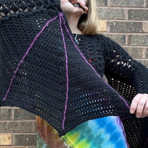 May include: Black crochet top with a bat wing design. The top has a large open stitch pattern and is accented with purple yarn.