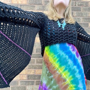 May include: A black crochet top with bat wings. The sleeves are long and have a purple trim. The top is cropped and has a loose fit.