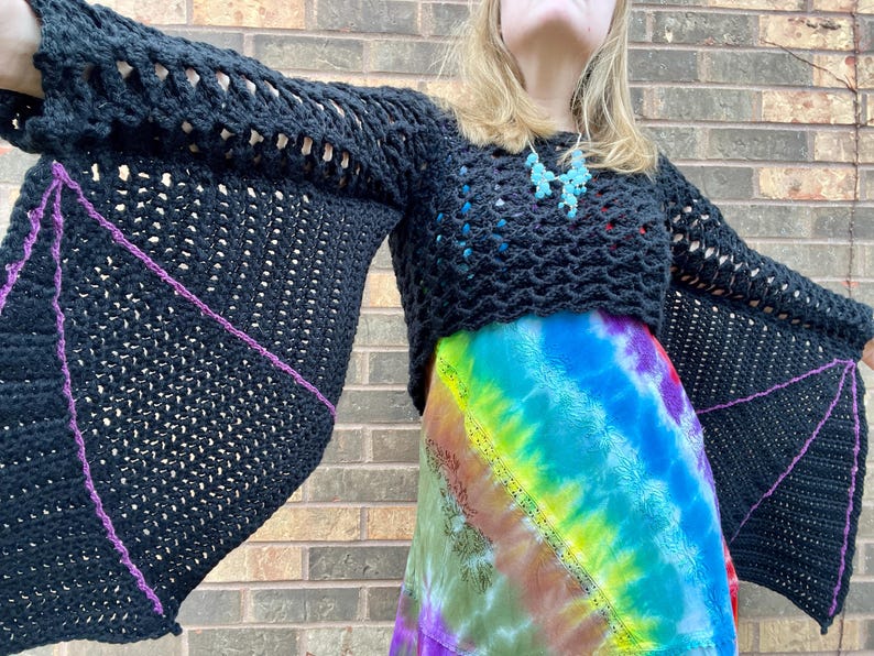 May include: A black crochet top with bat wings. The sleeves are long and have a purple trim. The top is cropped and has a loose fit.