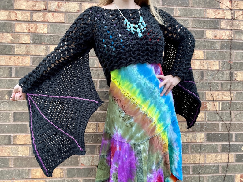 May include: Black crochet top with bat wing sleeves. The sleeves are lined with purple yarn. The top is cropped and has a loose fit.