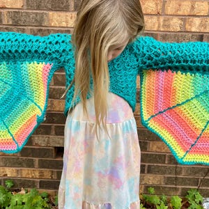 May include: A teal crochet top with rainbow-colored wings. The top is cropped and has a loose fit. The wings are made of a crocheted fabric with a gradient of colors.