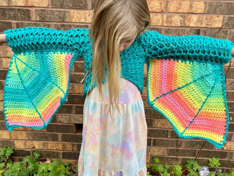 May include: A teal crochet top with rainbow-colored wings. The top is cropped and has a loose fit. The wings are made of a crocheted fabric with a gradient of colors.