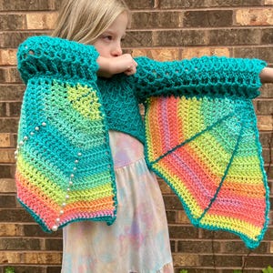 May include: A teal crochet top with rainbow ombre sleeves and a matching rainbow ombre crochet wing. The top is cropped and the wings are attached to the sleeves. The wings are decorated with white beads.