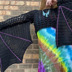 May include: A black crochet top with bat-shaped sleeves. The sleeves have a purple trim and are crocheted in a delicate pattern. The top is cropped and is worn over a tie-dye skirt.