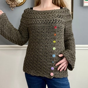 Könnte beinhalten: Ein grüner Häkelpullover mit Glockenärmeln und einem Regenbogen an Knöpfen an der Seite.