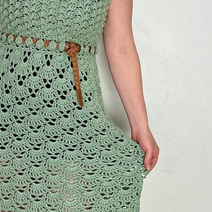 May include: A sage green crochet dress with a brown leather belt and beaded accents. The dress has a fitted bodice and a flowing skirt.