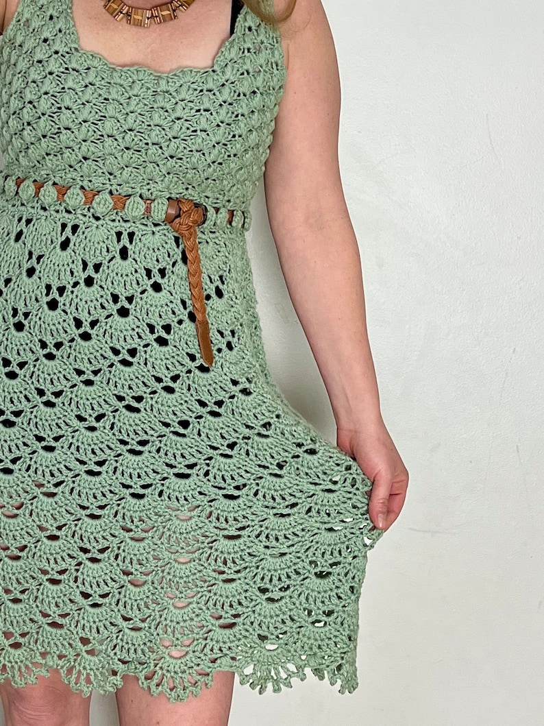 May include: A sage green crochet dress with a brown leather belt and beaded accents. The dress has a fitted bodice and a flowing skirt.
