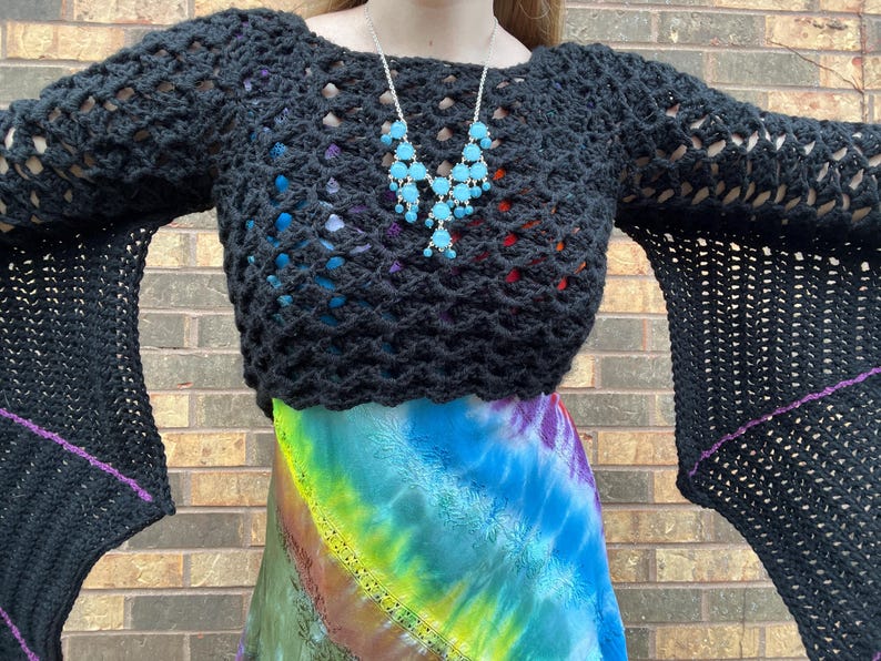 May include: A black crochet top with long bell sleeves and a tie-dye skirt. The top has a loose fit and a wide neckline. The skirt is a rainbow tie-dye pattern with a long, flowing silhouette.