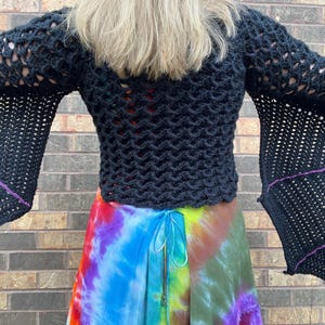 May include: Black crochet top with long bell sleeves and a purple trim. The top is worn over a colorful tie-dye skirt.