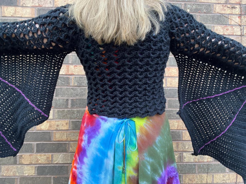 May include: Black crochet top with long bell sleeves and a purple trim. The top is worn over a colorful tie-dye skirt.