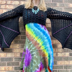 May include: A black crochet top with bat-like sleeves. The sleeves are lined with a purple trim. The top is cropped and worn over a tie-dye skirt.