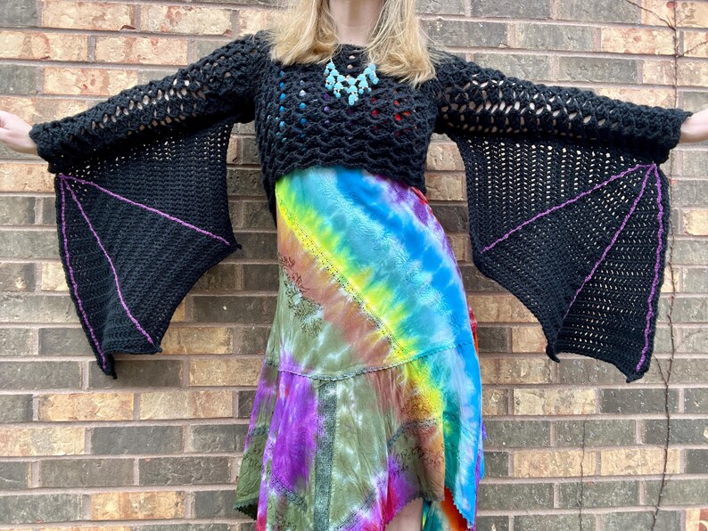 May include: A black crochet top with bat-like sleeves. The sleeves are lined with a purple trim. The top is cropped and worn over a tie-dye skirt.