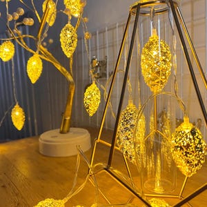 May include: A set of gold-toned pinecone-shaped LED string lights. Some lights are draped over a gold-coloured tree-shaped lamp with white pearl accents, while others are suspended inside a geometric gold metal frame with a glass cylinder.