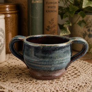 May include: A handcrafted ceramic bowl with two handles, glazed in shades of blue and brown. The bowl has a rustic, textured appearance, perfect for serving soups or stews. The bowl is sitting on a beige crocheted doily.