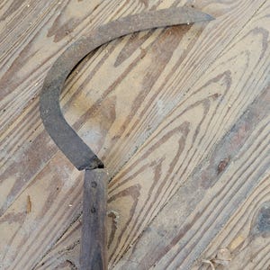 Puede incluir: Una hoz de metal oxidado y curvado con un mango de madera. La hoja en forma de media luna parece desgastada. El mango es de color marrón oscuro y cilíndrico. La herramienta está sobre una superficie de madera envejecida.