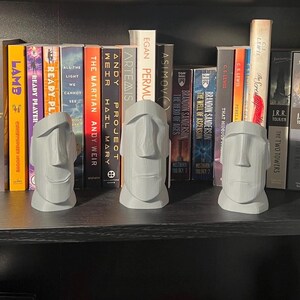 May include: Three grey stone-like bookends shaped like the heads of the Easter Island statues. The bookends are on a black bookshelf with a variety of books behind them.