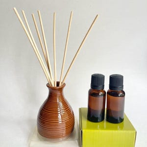 Puede incluir: Un jarrón difusor de cerámica marrón con estrías horizontales que contiene varias varillas de madera clara, acompañado de dos frascos pequeños de vidrio ámbar con tapas negras sobre una caja de color verde lima.