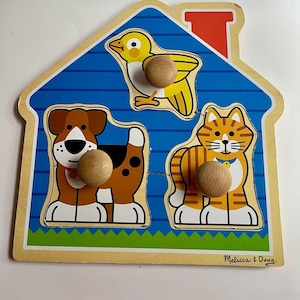 May include: A wooden house-shaped peg puzzle featuring three removable pieces: a yellow bird, a brown and white dog, and an orange tabby cat. Each animal piece has a round wooden knob for easy grasping. The base displays a blue house design.