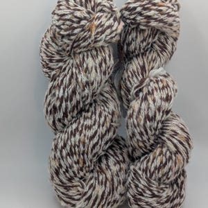 May include: Two skeins of yarn in a brown, cream, and tan speckled pattern. The yarn is tightly wound, showing the texture and color variations. Ideal for knitting or crocheting projects.