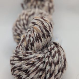 May include: Close-up of a skein of yarn with a textured appearance. The yarn is a mix of brown, cream, and peach colors, creating a speckled effect. The yarn is tightly wound, suggesting it is ready for use in knitting or crochet projects.