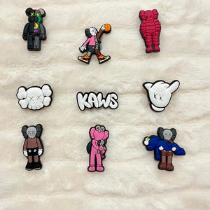 May include: A selection of colourful KAWS-themed shoe charms. The charms feature various KAWS character designs in black, red, pink, and white, along with the KAWS logo. These charms are designed to be attached to shoes.