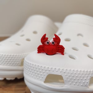 May include: A pair of white clogs with a red crab-shaped shoe charm. The charm is centered on the top of one shoe. The clogs have multiple holes and a textured surface.