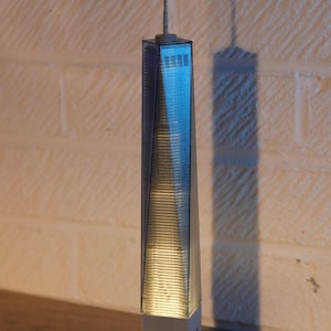 May include: A clear plastic model of the Freedom Tower, also known as One World Trade Center, on a wooden base. The model is lit from within, creating a warm glow.
